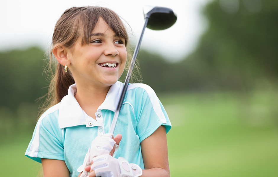 School-aged girl holding golf club and wearing golf attire and glove, smiling